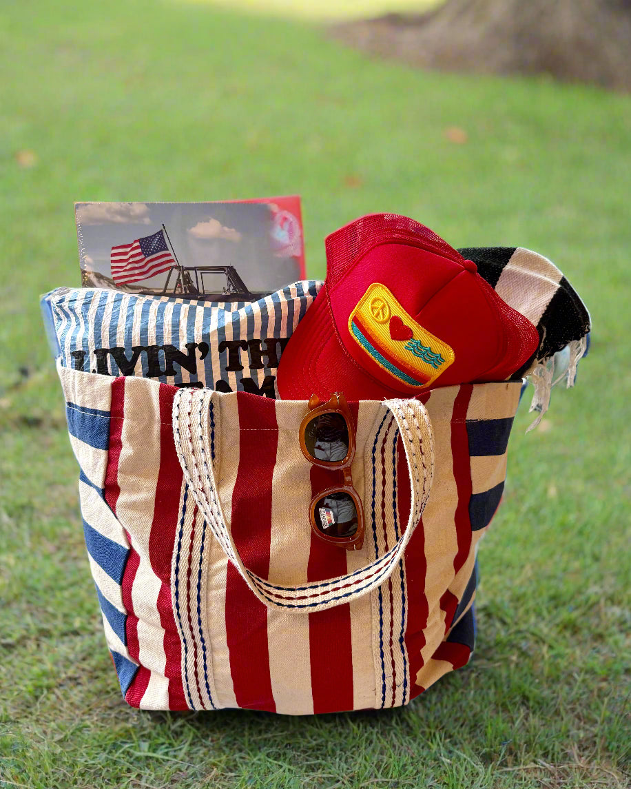 The DEBBIE KATZ Cabana Beach Bag, in red, white, and blue stripes, holds a towel, red cap, sunglasses, and a roller coaster magazine atop grass—ideal for summer picnics or beach parties.