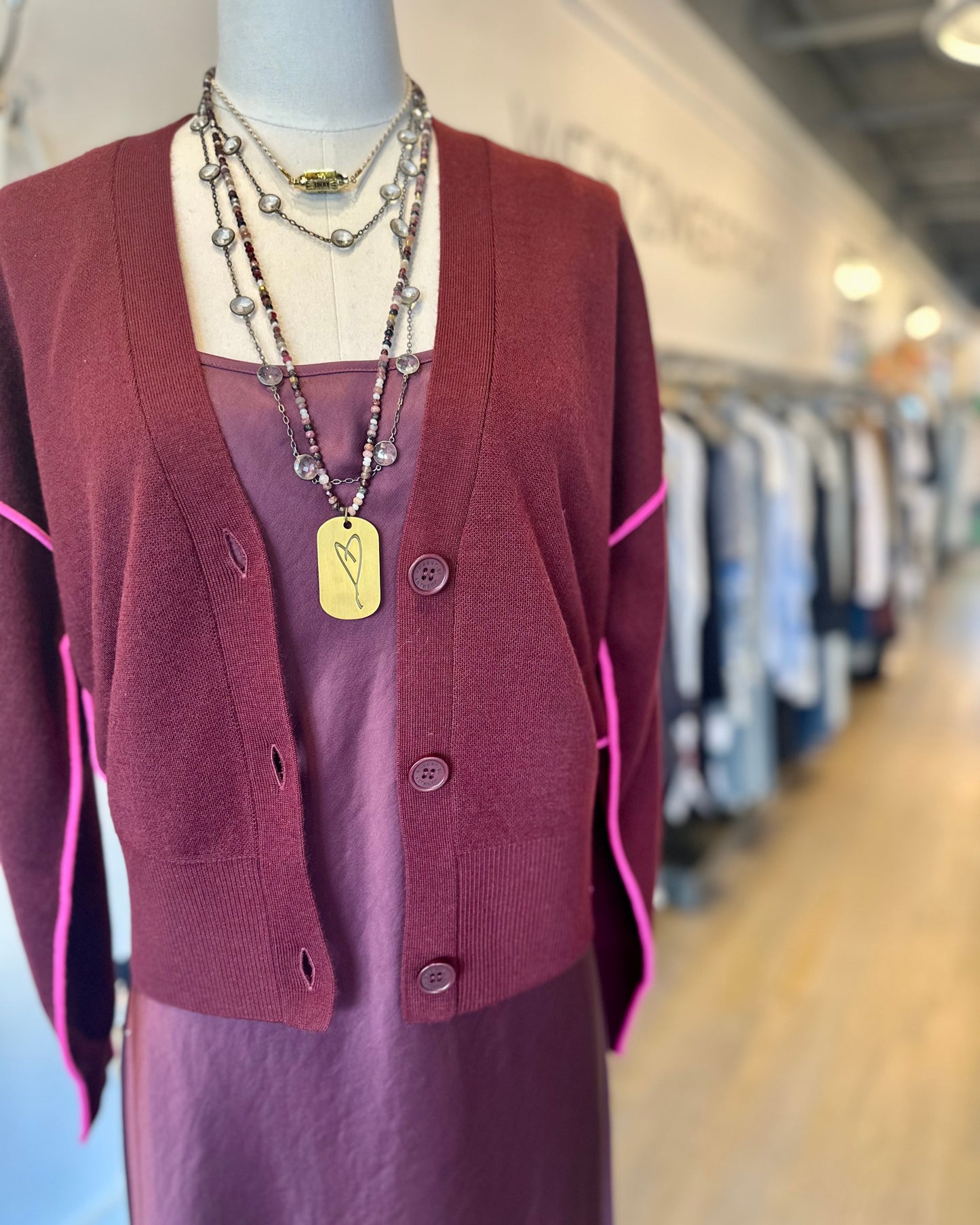A mannequin in the Ileo Cardigan by Essentiel Antwerp, a mid-weight burgundy knit with contrast piping, layered over a silky dark mauve dress and necklaces, stands in a boutique with blurred racks behind.