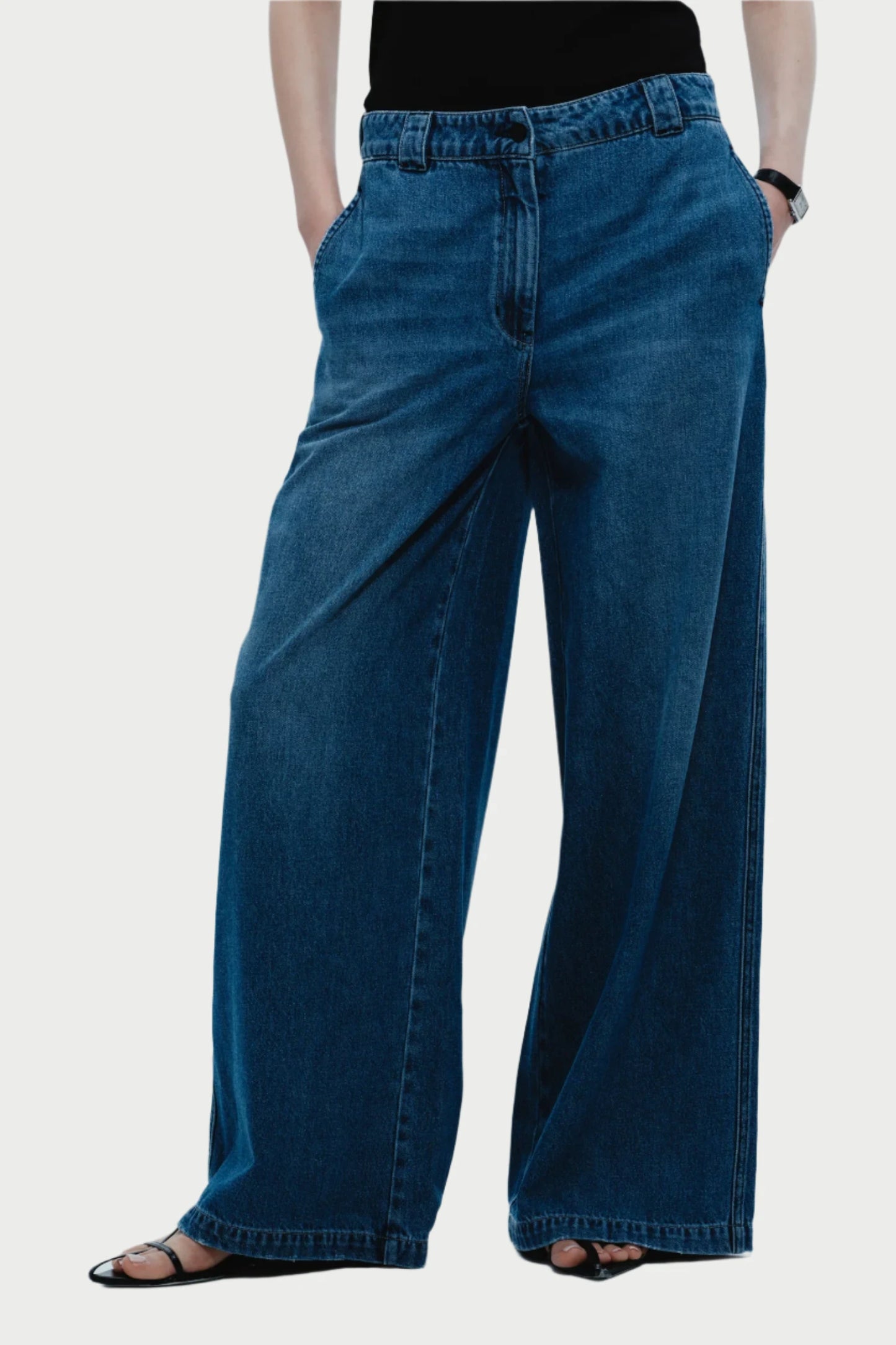 Model wears Saint Art NY’s Amber Wide Leg Denim Pant in Tencel denim, paired with a black top, sandals, and watch, standing with hands in pockets against a plain white background.