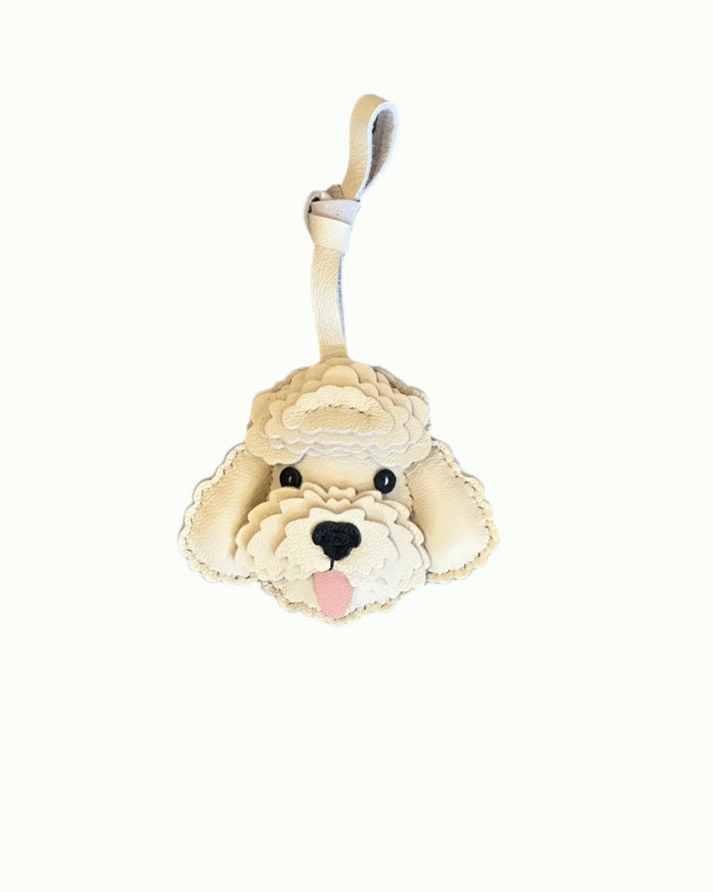 The Charm XO Leather Poodle Bag Charm features a small white poodle with floppy ears, black nose, and pink tongue—an adorable accessory or keychain, pictured on a wood floor.