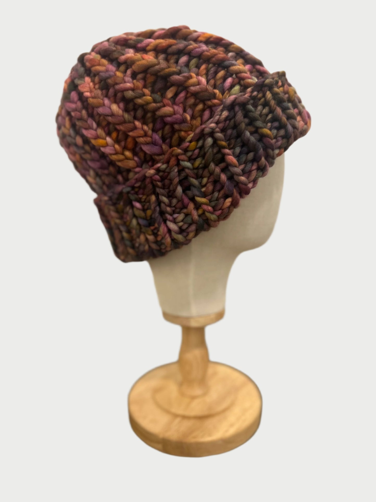 The Soft Wool Beanie by Meg Cohen, a multicolored, chunky knit made from soft wool, is showcased on a wooden stand. Lightweight yet warm, it features shades of purple, orange, and brown against a plain white background.