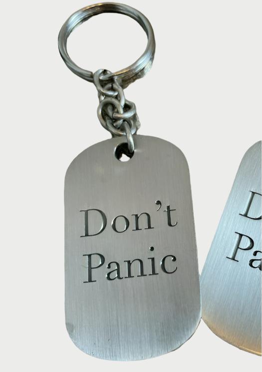 The Simply Eartha Don’t Panic Key Chain is a vintage-style silver metal tag engraved with "Don't Panic" in bold black letters, with another similar tag partially visible, enhancing its unique charm.