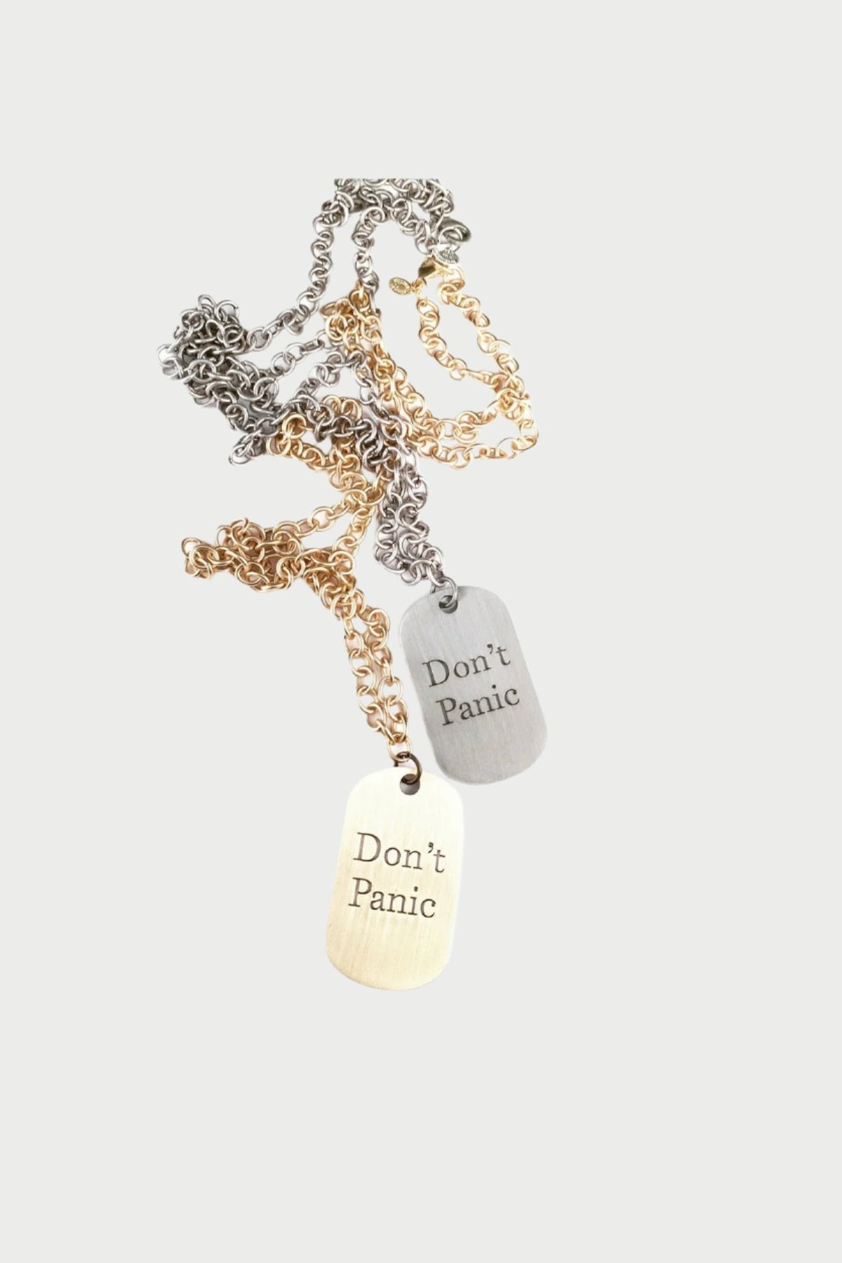A tangled collection of jewelry chains in silver and gold hues with two rectangular dog tags. Both tags have the engraved text "Don't Panic" on them. The tags are metallic, one in a gold color and the other in silver, showcased on heavy gauge chains for a striking look. The piece is called the Don't Panic Dog Tag Necklace by Simply Eartha.