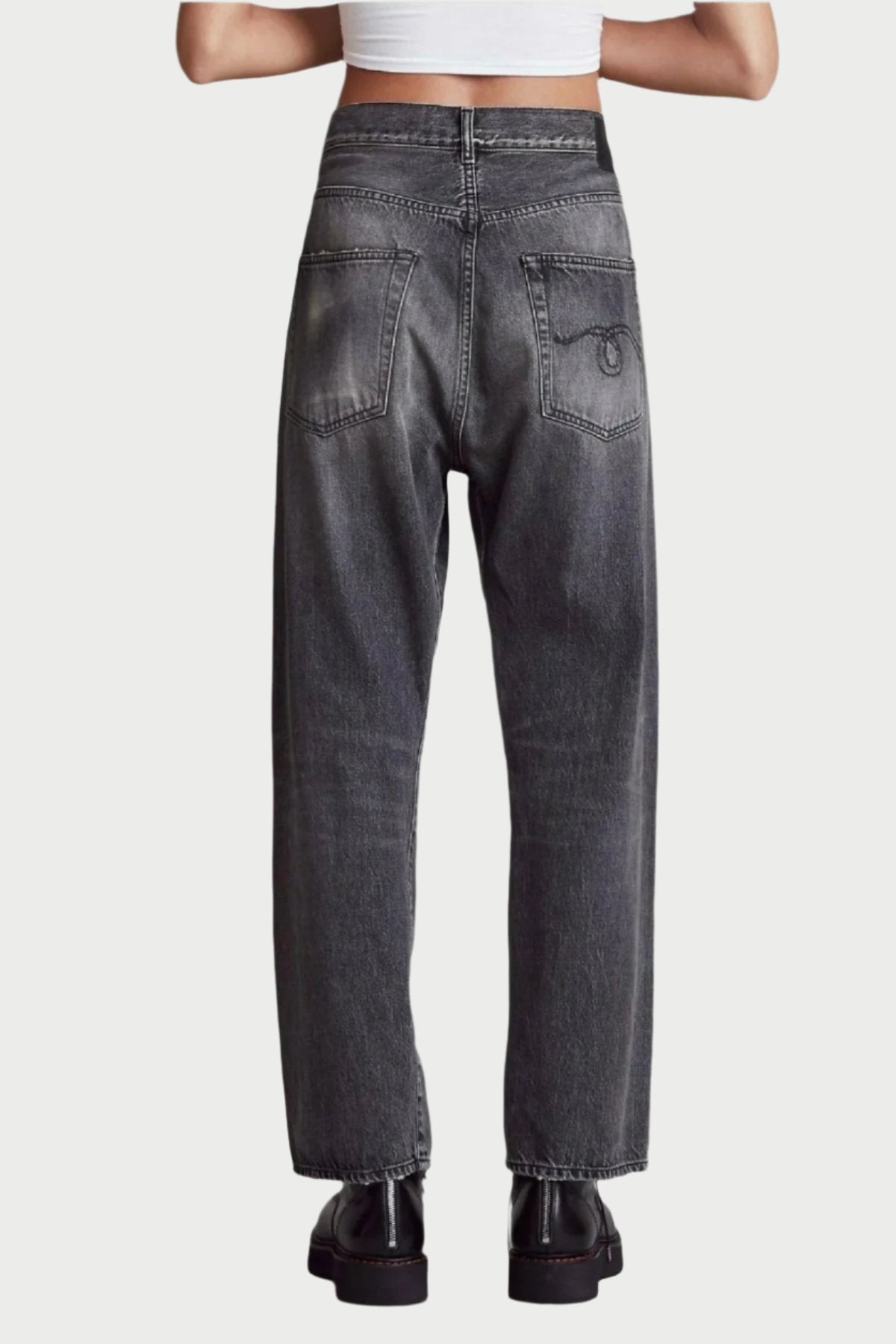 A person is shown from the back wearing the coolest jeans: high-waisted, faded black R13 Crossover Jean in Leyton Black paired with chunky black shoes. They also sport a white crop top against a plain gray background. For the perfect fit, consider sizing down.
