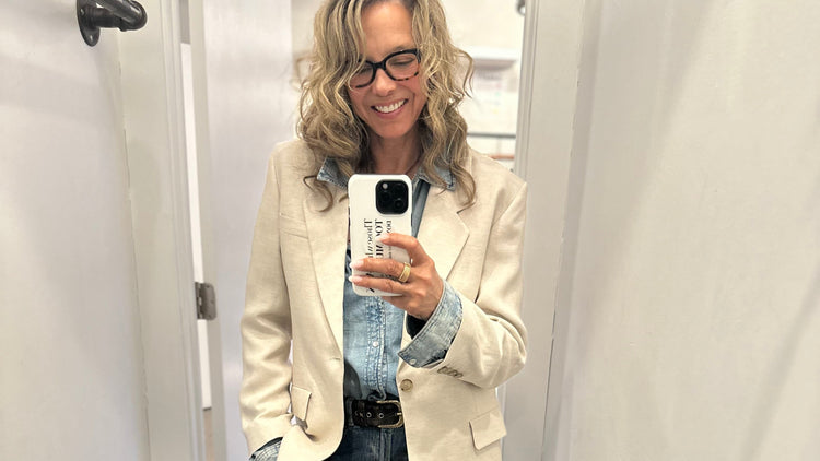 A person with wavy hair wearing glasses, a beige blazer over a denim shirt and jeans, poses for a mirror selfie. They're standing in a well-lit room and smiling, holding a phone with a case.