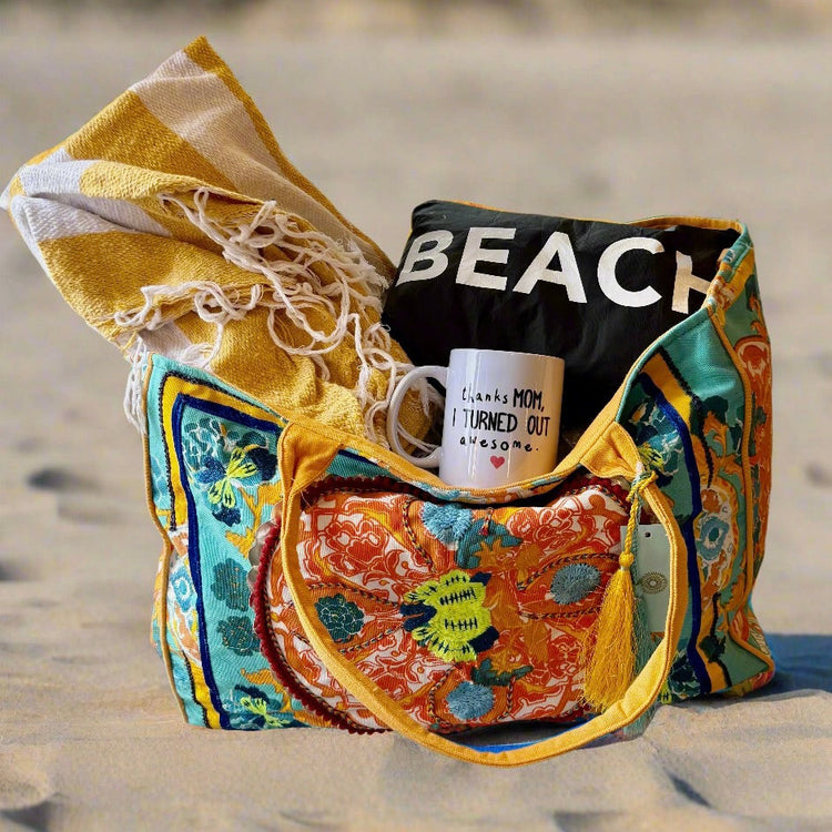 A colorful beach bag sits on sand, filled with a yellow striped towel, a black "BEACH" shirt, and a white mug that reads, "Thanks Mom, I turned out awesome.