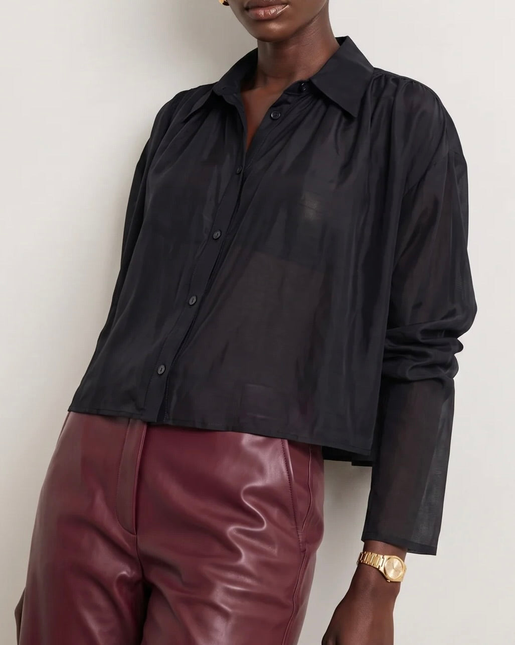 A person wearing The Shirt Hayden Shirt Black, a long-sleeved cropped button-up with a collar, paired with dark red trousers.