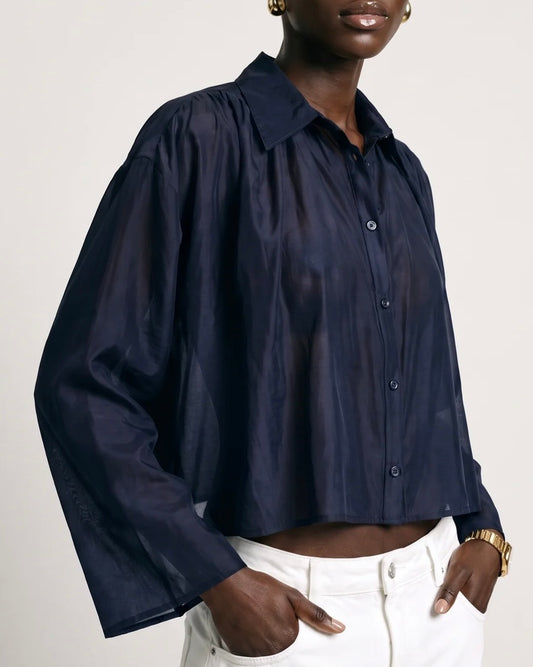 A person wears The Shirt Hayden Shirt Navy, a cropped, semi-sheer button-up with a relaxed fit and long sleeves, paired with white trousers.