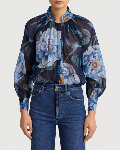 A navy blue Smythe Gathered Blouse Navy Floral with a high neckline, long sleeves, and a large blue floral print, worn tucked into blue jeans.