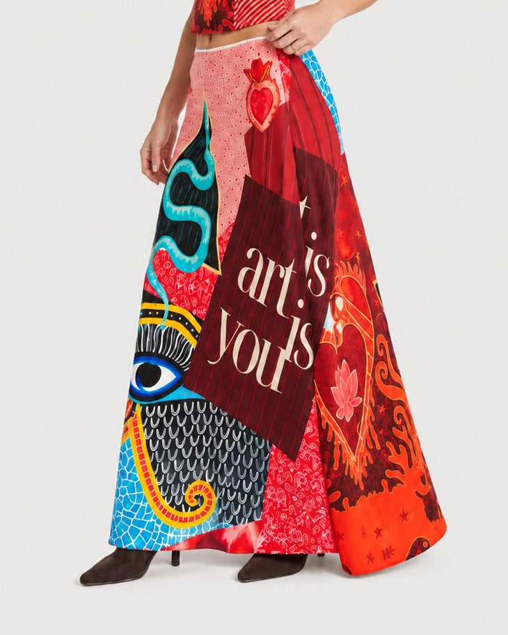 ME369 Blakely Slip Skirt Red Aura, a bias-cut maxi skirt featuring a multicolored collage of graphic patterns, including an eye, a sacred heart, and the text “art is yours.”