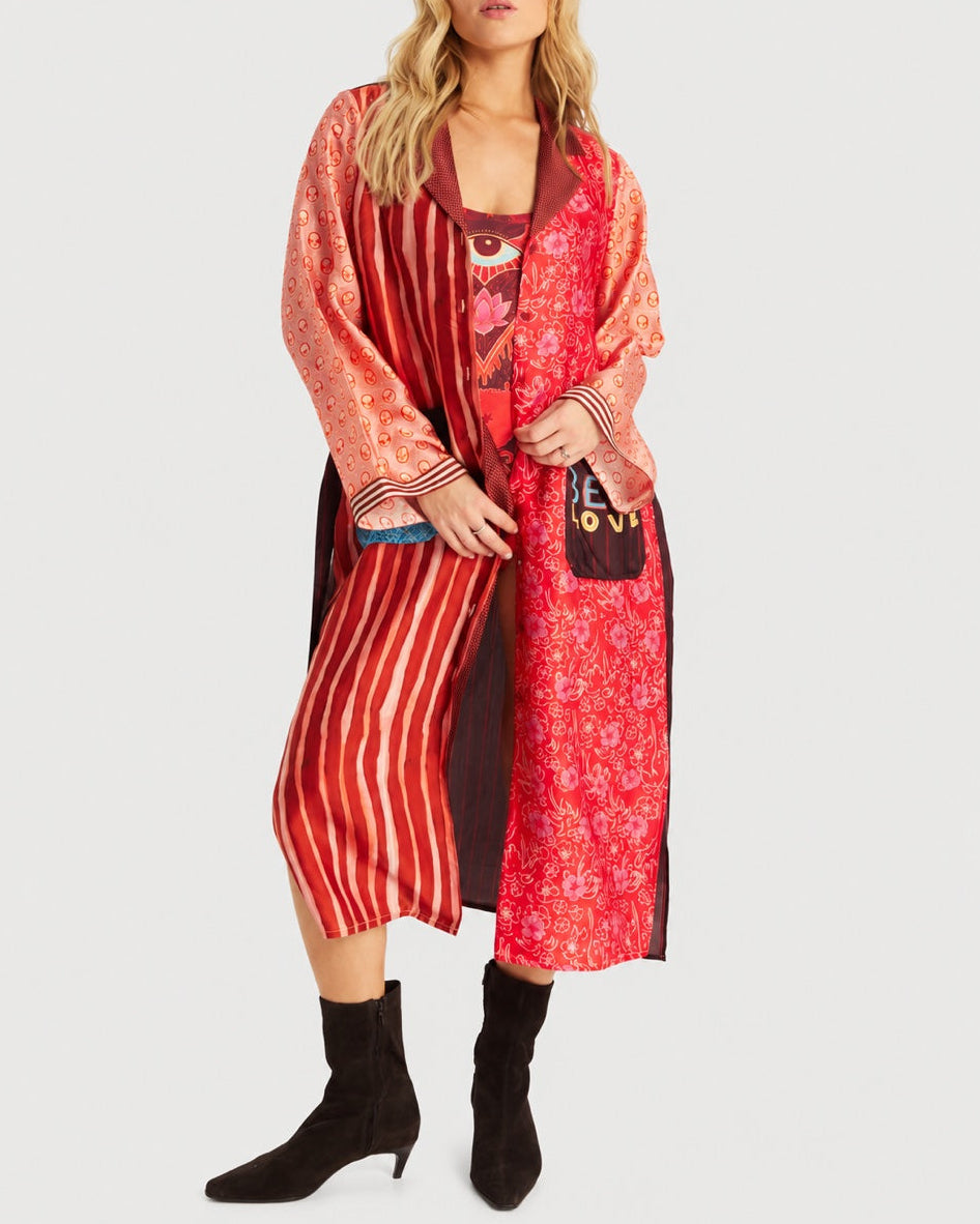 A person wears a red ME369 Nova Kimono Dress Red Aura featuring a mix of vertical stripes, floral patterns, and a graphic of an eye on the chest, paired with black ankle boots.