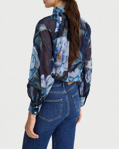 Rear view of a person wearing a Smythe Gathered Blouse Navy Floral with a high neck, light blue floral print, long gathered sleeves, and dark blue jeans.