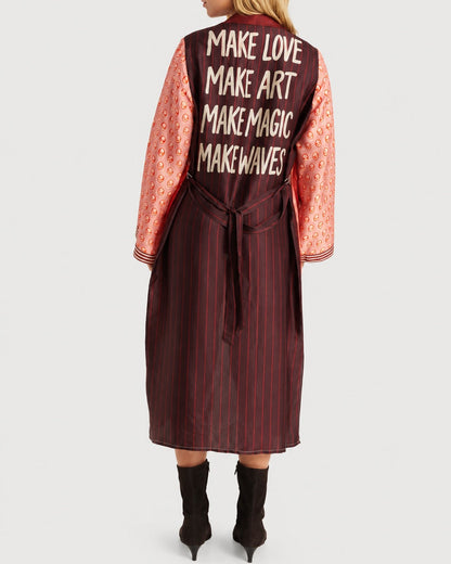 Back view of a person wearing a red pinstriped ME369 Nova Kimono Dress Red Aura with pink sleeves featuring a repeating pattern and the text “MAKE LOVE, MAKE ART, MAKE MAGIC, MAKE WAVES” printed on the back.