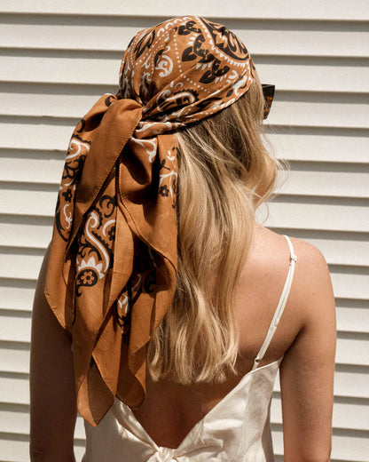 A woman with long blonde hair, seen from behind, wears the Maison Lecomte Flament Bandana Scarf Camel tied around her head. She stands before a white, horizontal slatted wall in a sleeveless white top.