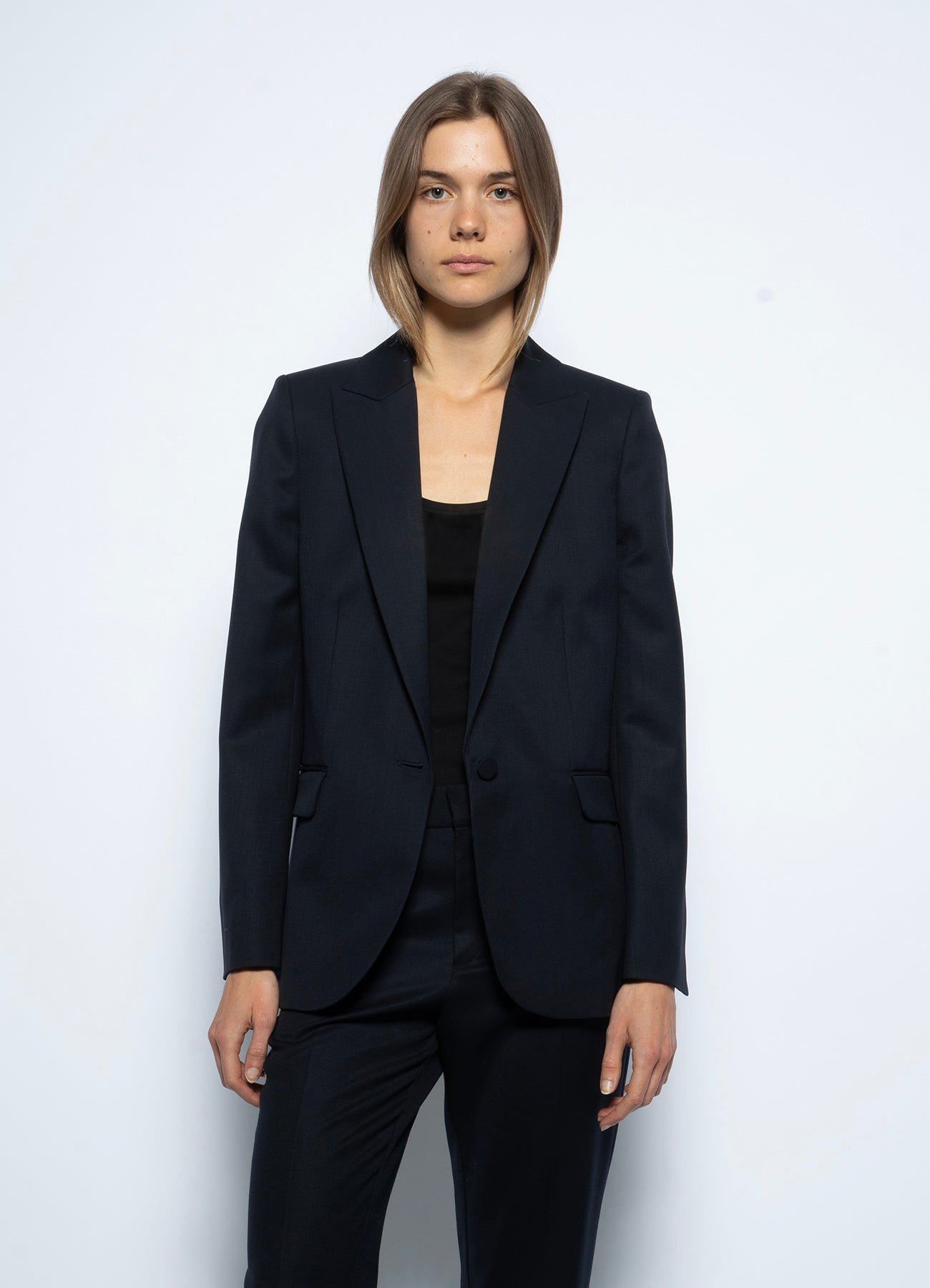 A woman with straight, shoulder-length hair wears the Zadig & Voltaire Tailored Navy Blazer over a black top and matching pants, standing against a plain light background and looking directly at the camera with a neutral expression.