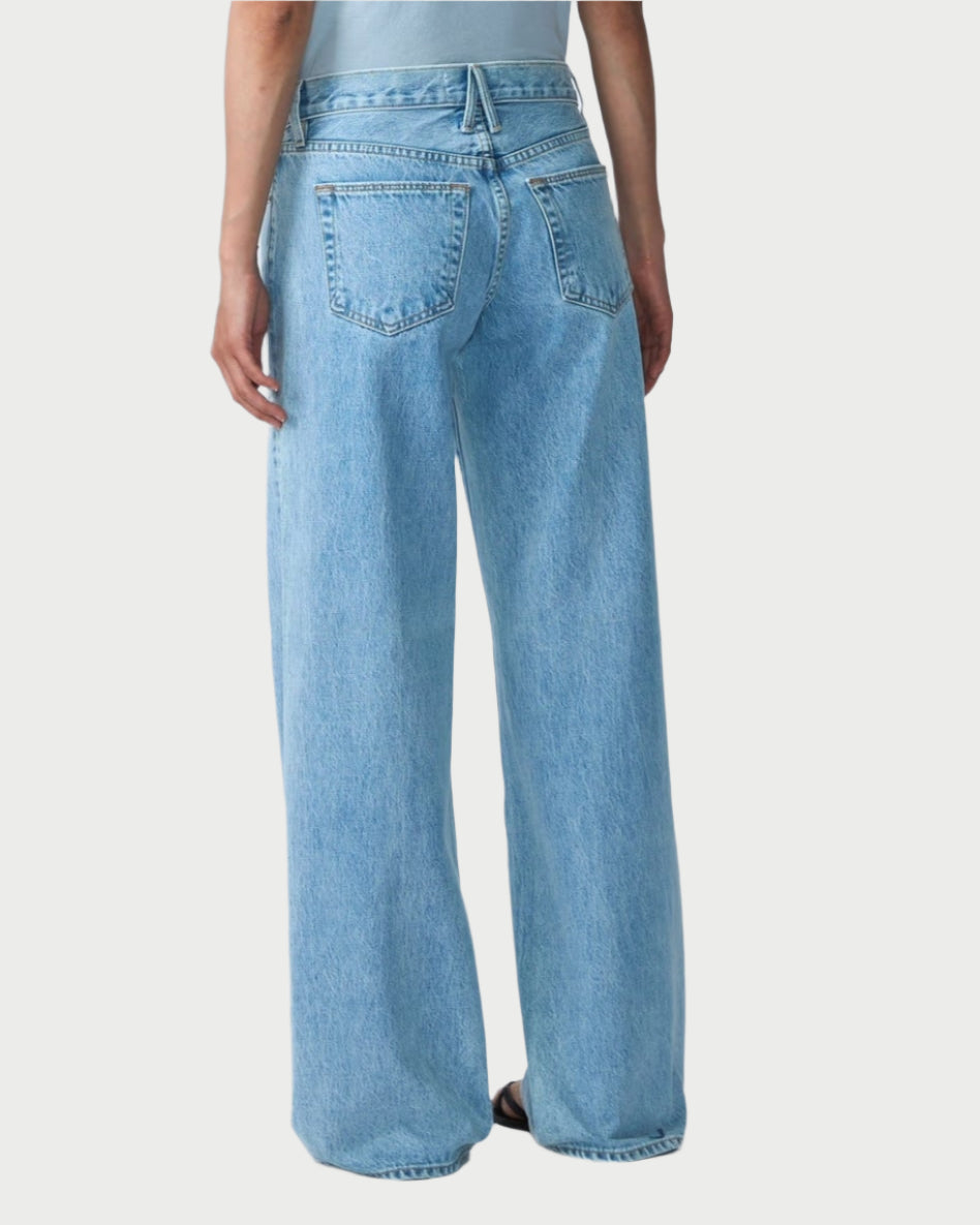 A person with long, dark hair stands against a plain white background wearing a light gray T-shirt and loose-fitting SLVRLAKE's Mica Jeans in Clear Skies. The relaxed silhouette exudes a vintage-inspired vibe, while the fabric tells stories of regenerative cotton.