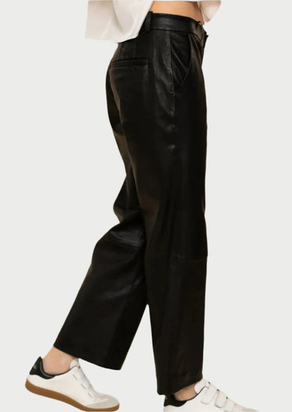 A person is wearing SPRWMN's Cropped Baggy Leather Trousers in black, paired with white sneakers featuring brown soles. The photo captures a side view of the low-rise fit against a plain background, highlighting the sleek design of the lower body.