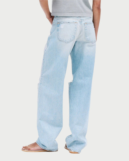 A person faces away, wearing a gray T-shirt and SLVRLAKE Tess Long Jeans in light blue with a curved leg silhouette and vintage-inspired finish. Their brown sandals complement the look, and their hair is tied up in a bun against a plain white background.