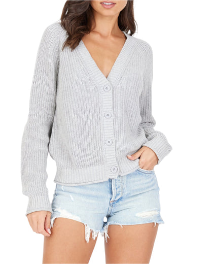 A woman with wavy brown hair is wearing a light gray, versatile Raleigh Cardigan from One Grey Day along with distressed denim shorts. She is standing with one hand in her pocket, posing confidently against a plain white background.