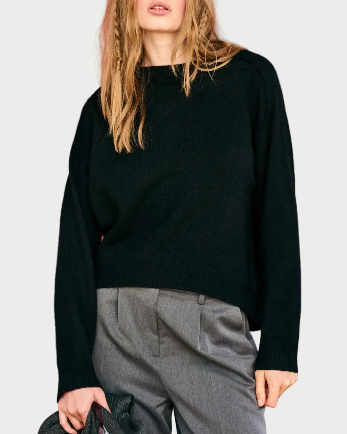 A person with long, light brown hair wears the JUMPER1234 Oversize Cashmere Crew in black and high-waisted gray trousers, holding a dark jacket against a plain white background.