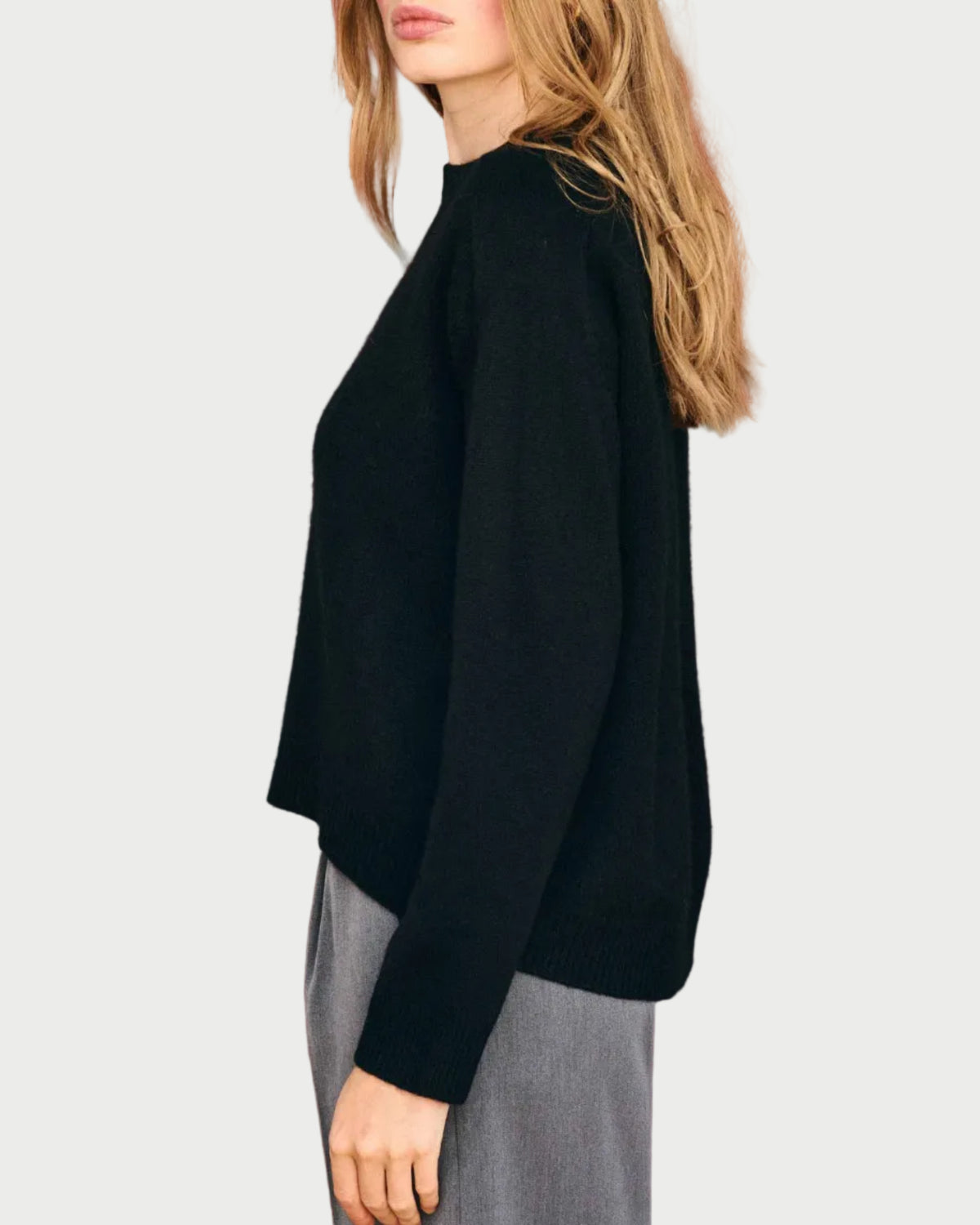 A woman with long, light brown hair stands sideways against a plain, light background, wearing the JUMPER1234 Oversize Cashmere Crew in black paired with gray pants.