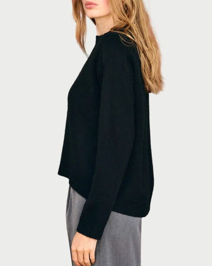 A woman with long, light brown hair stands sideways against a plain, light background, wearing the JUMPER1234 Oversize Cashmere Crew in black paired with gray pants.