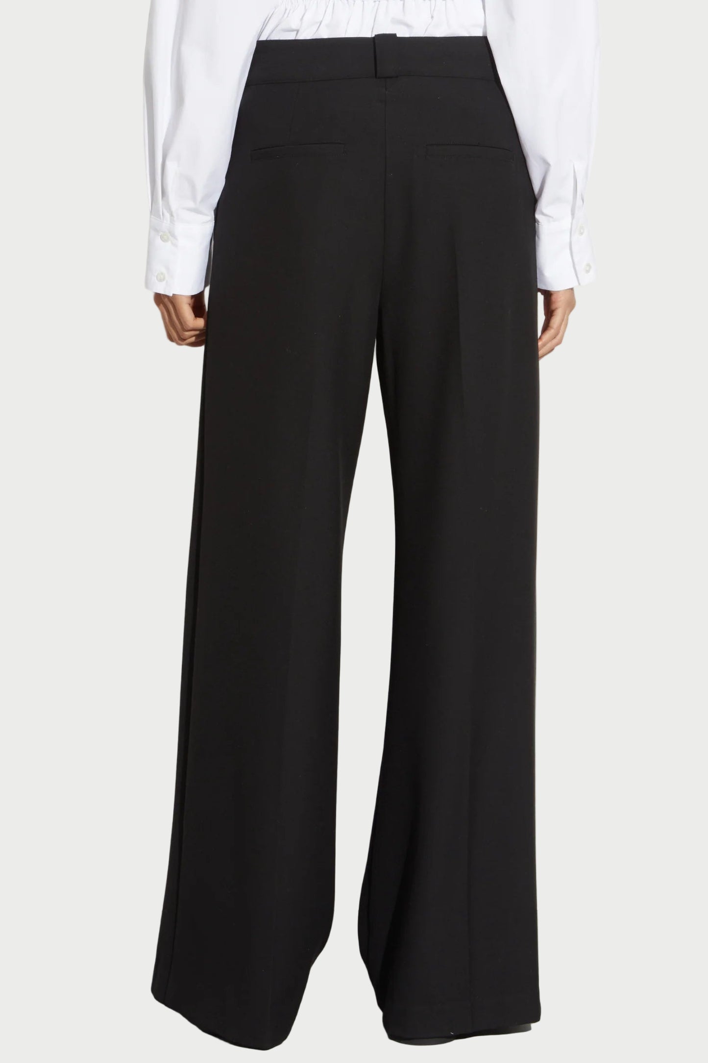 Someone wearing Saint Art NY’s Tiffany Wide Leg Pants in black and a white long-sleeve shirt, seen from the back against a plain background.