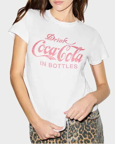A woman wearing a white short-sleeve graphic T-shirt with a pink distressed Coca-Cola logo and text reading 'Drink Coca-Cola IN BOTTLES,' paired with leopard-print pants against a plain white background.