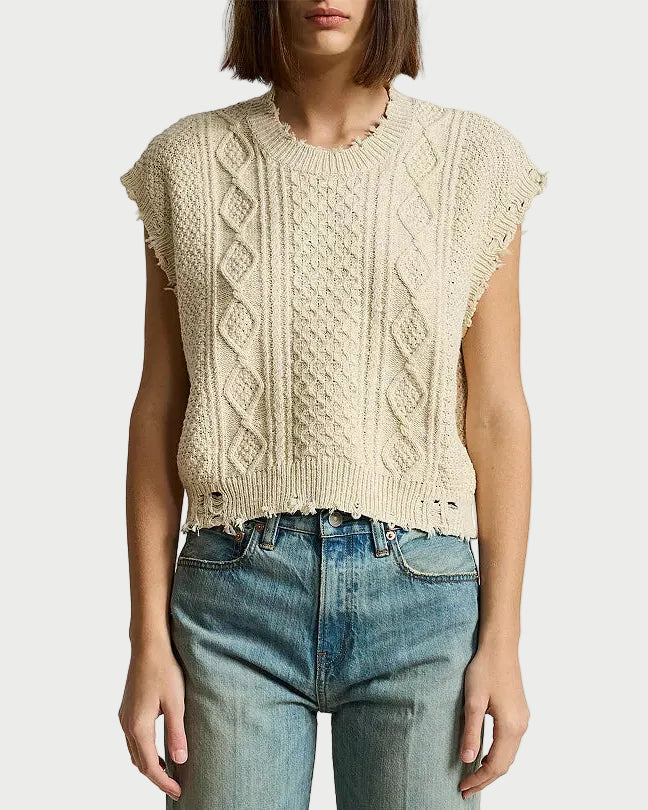A person wearing the Denimist DENIMIST Boyfriend Cable Vest—a sleeveless, beige cable-knit crewneck sweater vest with frayed edges—paired with high-waisted blue jeans. The background is plain white, and the person’s face is partially out of frame.