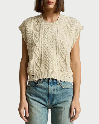 A person wearing the Denimist DENIMIST Boyfriend Cable Vest—a sleeveless, beige cable-knit crewneck sweater vest with frayed edges—paired with high-waisted blue jeans. The background is plain white, and the person’s face is partially out of frame.