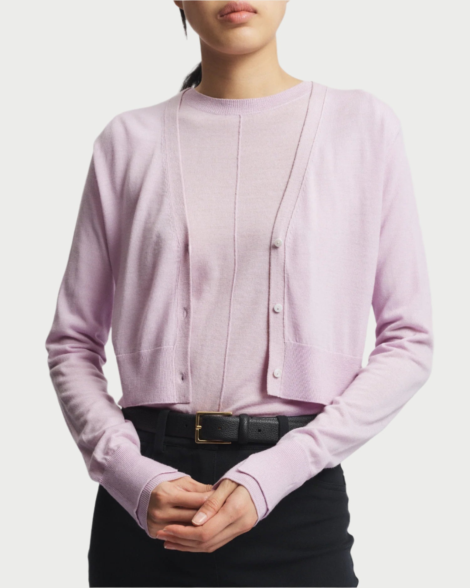 Elegantly dressed in a Saint Art NY Nixon Cropped Cardigan made of luxury knitwear in light purple over a matching top, the person wears a black belt and dark pants. Their hands are clasped against a white background.