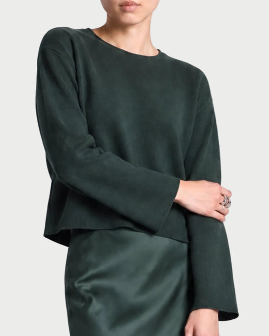 A person wears the Saint Art Marley Double Knit Crewneck by Saint Art NY in dark green with a matching satin skirt, one arm crossed and a silver ring on their finger, against a plain light gray background.