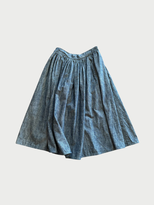 The 176 Indigo Skirt by A Friend Made is a unique, knee-length, high-waisted blue chambray midi with a gathered flared silhouette, shown on a white background.