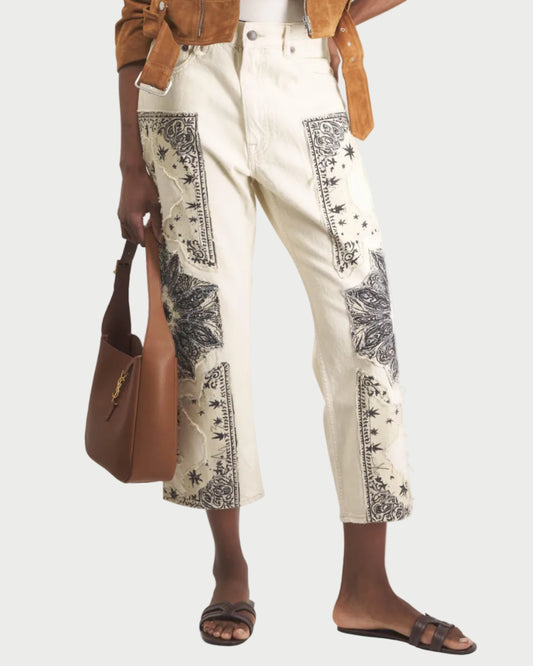 Person wearing a brown jacket and white pants with black patterns, holding a brown bag on a white background.