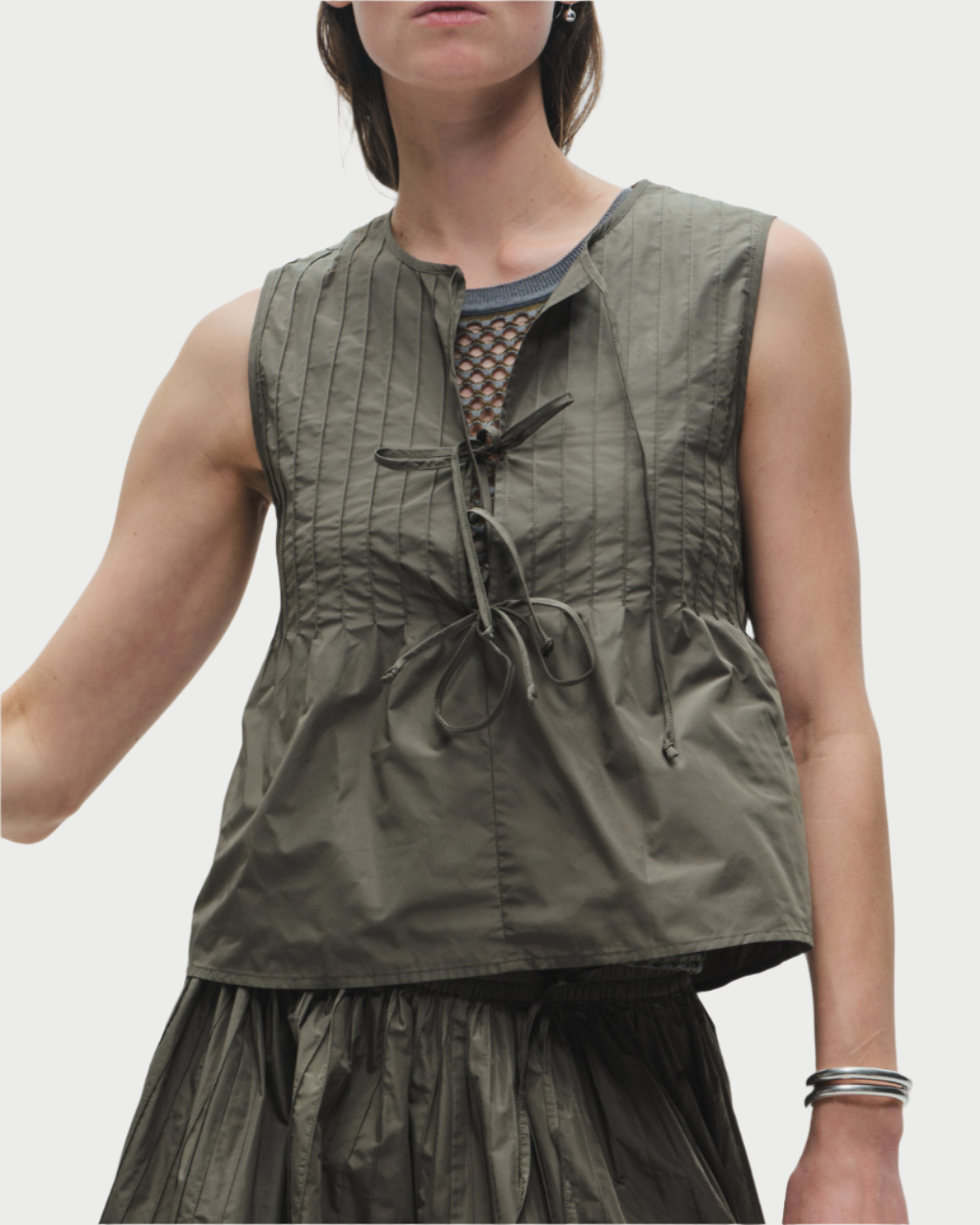 A person wears the sleeveless Saint Art NY Nina Tie Front Top in olive, featuring drawstrings and a pleated, slightly crinkled texture, paired with a matching skirt against a light plain background.