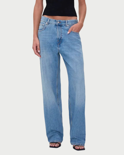 Person wearing a black sleeveless top and blue jeans standing against a beige curtain backdrop.