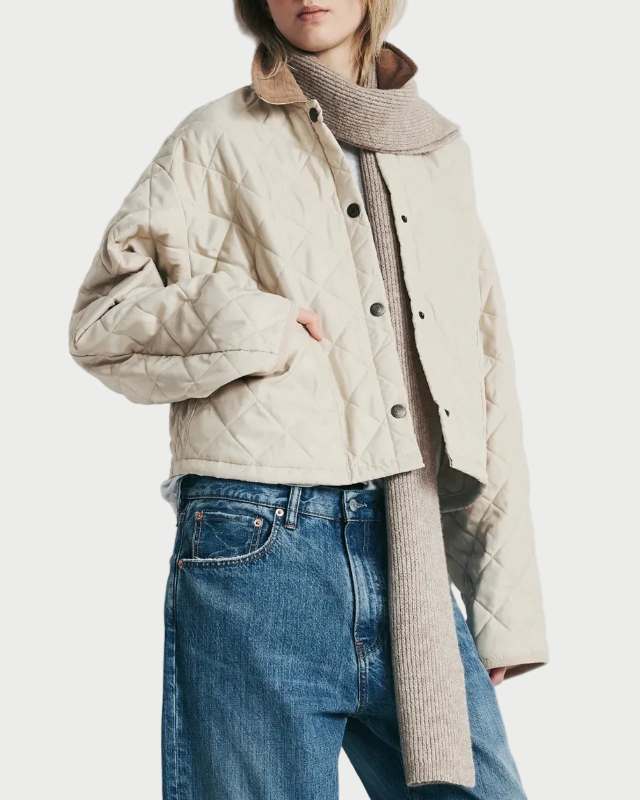 A model wearing a beige quilted cropped jacket with a high collar, styled with a ribbed beige turtleneck, blue jeans, and sunglasses against a plain light gray wall.
