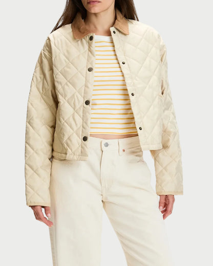 A model wearing a cream quilted jacket with a brown collar, paired with cream wide-leg pants, a yellow and white striped top, and tan mule shoes, set against a plain white studio background.