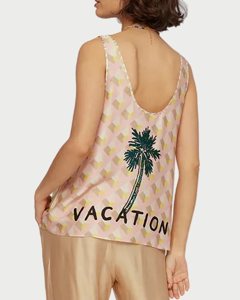 Woman wearing a sleeveless top with 'Vacation' text and palm tree design, paired with gold pants, against a beach backdrop.