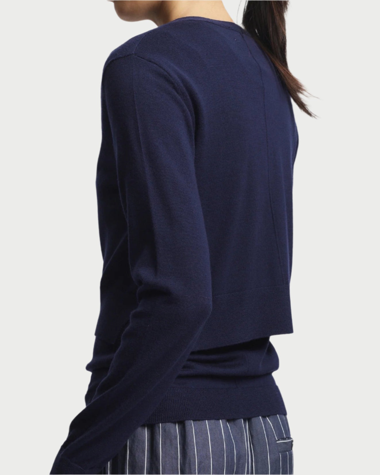 A person with long dark hair is shown from the back, wearing a cozy Nixon Cropped Cardigan by Saint Art NY and gray pants featuring white vertical stripes. The backdrop is plain white.