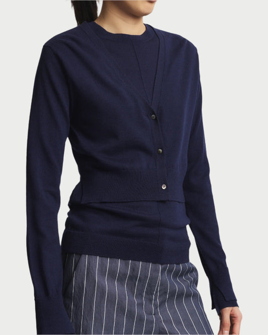 A person in a Nixon cropped Cardigan from Saint Art NY (dark blue) over a matching shirt and blue pinstripe pants stands chicly against a white background. The cardigan's button-up design enhances versatility and style, maintaining a casual look.