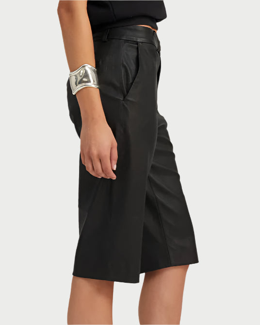 A person is dressed in black Bermuda trouser shorts from SPRWMN, a matching top, and a large silver cuff bracelet on the right wrist while standing against a plain light gray background.
