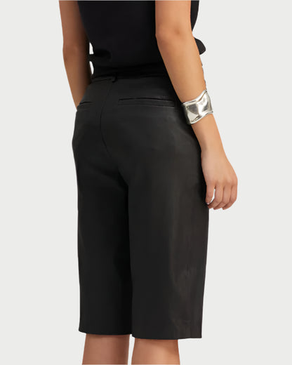 From behind, a person is wearing a black top with SPRWMN's knee-length Bermuda Trouser Shorts. A wide silver bracelet graces their left wrist, while the French stretch-leather from the shorts adds elegance against the light-colored background.