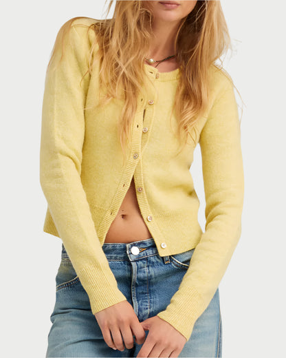 A person with long, loose hair stands against a light background, wearing SPRWMN's Baby Cashmere Cardigan in yellow. Crafted from sustainably grown materials, this cardigan is partially unbuttoned over a white top and paired with blue jeans.