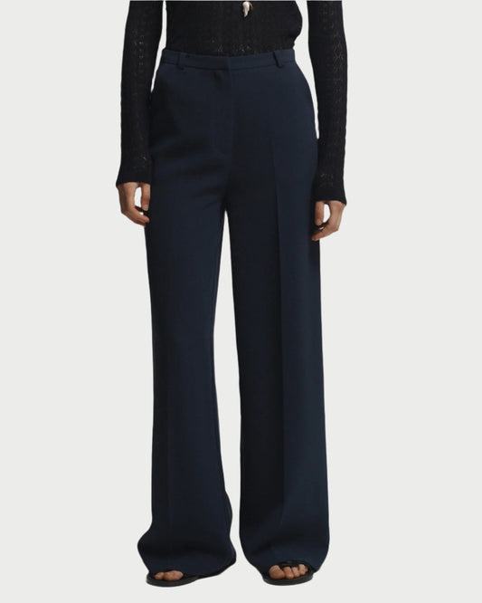 A person stands against a plain white background, hands relaxed by their sides, wearing Saint Art NY's Rhodes Wide Leg Pant in navy crepe with a back waistband buckle and a black long-sleeve top.