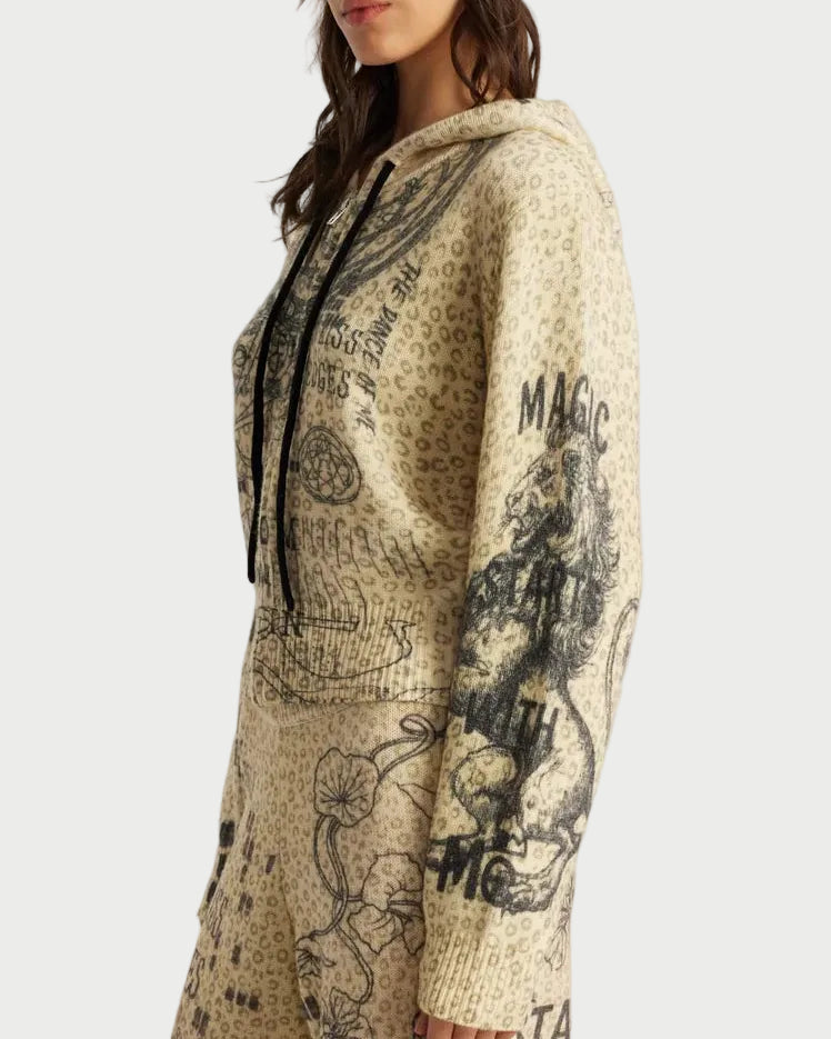 A woman wearing a beige and black graphic print hooded sweater and matching beanie, photographed against a plain light gray studio background.