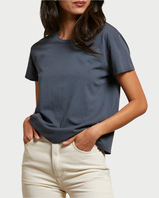 A woman stands with hands in pockets against a light background, wearing the perfectwhitetee Harley Boxy Crew Night—a dark gray, 100% cotton relaxed-fit tee—and high-waisted light beige pants. Her head is slightly tilted and her dark hair is loose.