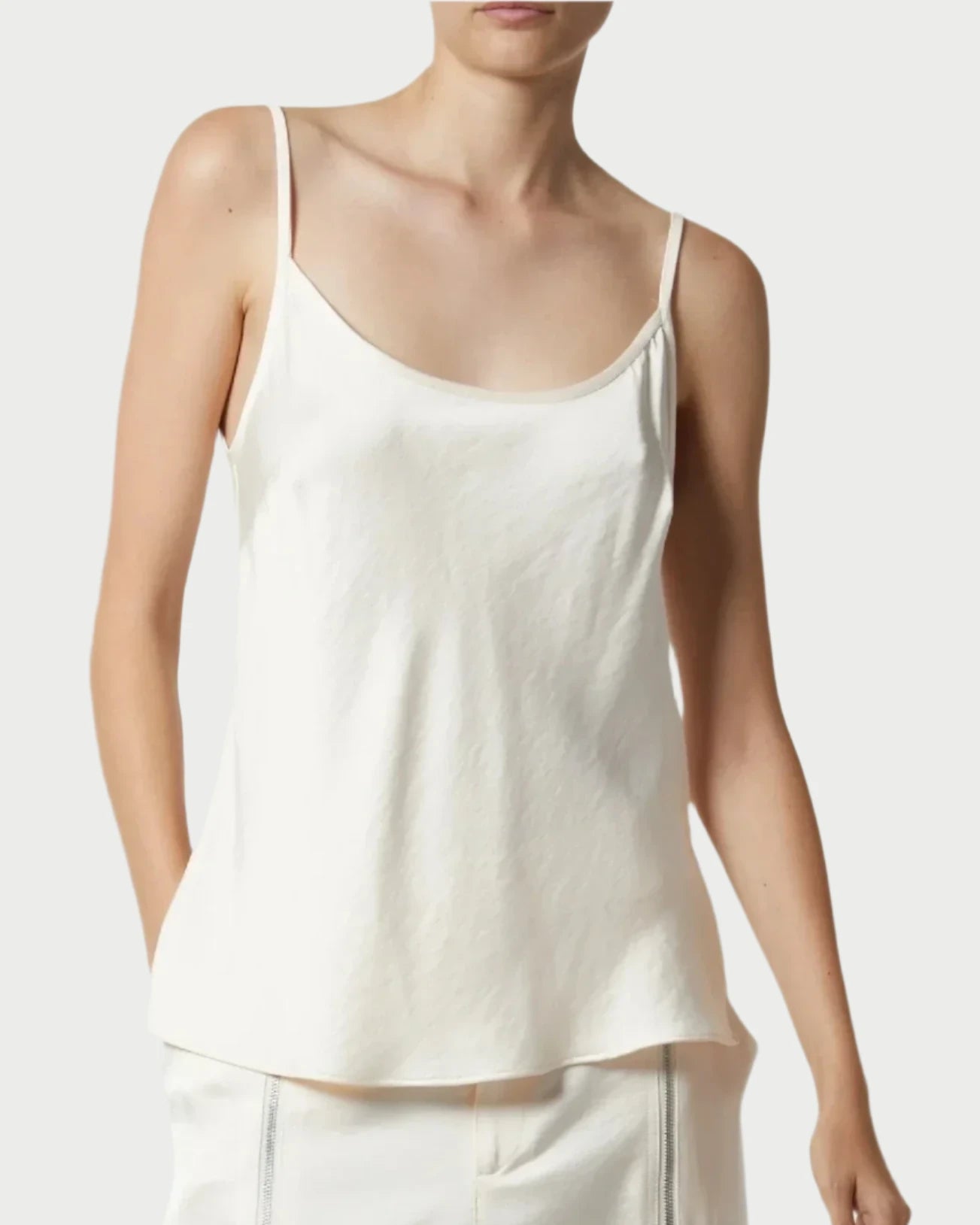 A person wearing a Saint Art NY Charmeuse Cami, a white, sleeveless camisole with thin straps, paired with white pants featuring zipper details. The silky-smooth fabric exudes modern femininity. Only the upper body is visible against a plain background, highlighting the simple, casual style of the outfit.