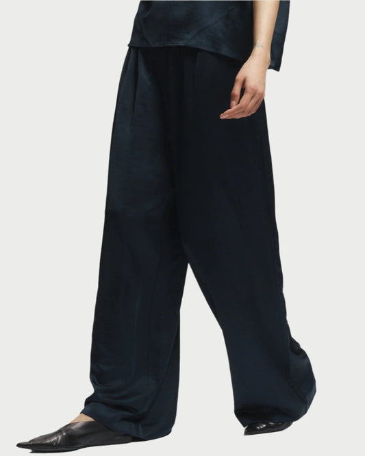 A person wears the Saint Art NY Paige Pull-On Pant—loose-fitting, dark navy, wrinkle-resistant with a ruched waistband—paired with pointed black shoes and a partial matching top, shown against a plain white background.