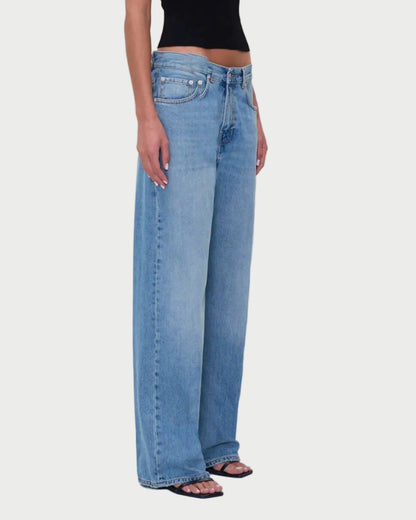 Person wearing a black sleeveless top and blue jeans standing in front of beige curtains.